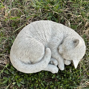 May include: A gray concrete garden statue of a cat curled up sleeping. The cat is in a curled position with its tail wrapped around its body. The statue is placed on a bed of green grass.c