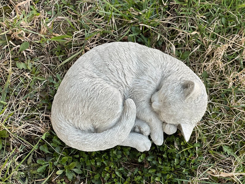 May include: A gray concrete garden statue of a cat curled up sleeping. The cat is in a curled position with its tail wrapped around its body. The statue is placed on a bed of green grass.c
