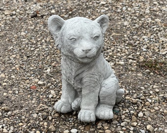 May include: A gray concrete statue of a tiger cub sitting with its paws crossed. The statue has a realistic appearance with detailed fur and facial features.