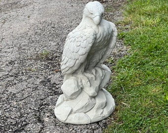 May include: A white concrete statue of an eagle perched on a rock. The eagle is facing to the right and has its wings folded. The statue is detailed with feathers and talons.