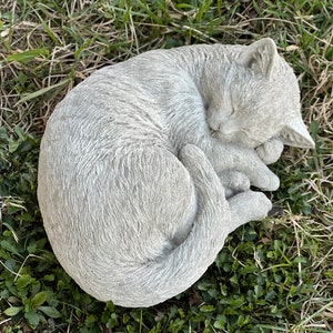 May include: A gray concrete garden statue of a cat curled up sleeping. The cat is lying on its side with its head resting on its paws. The statue is detailed with fur texture and realistic features.