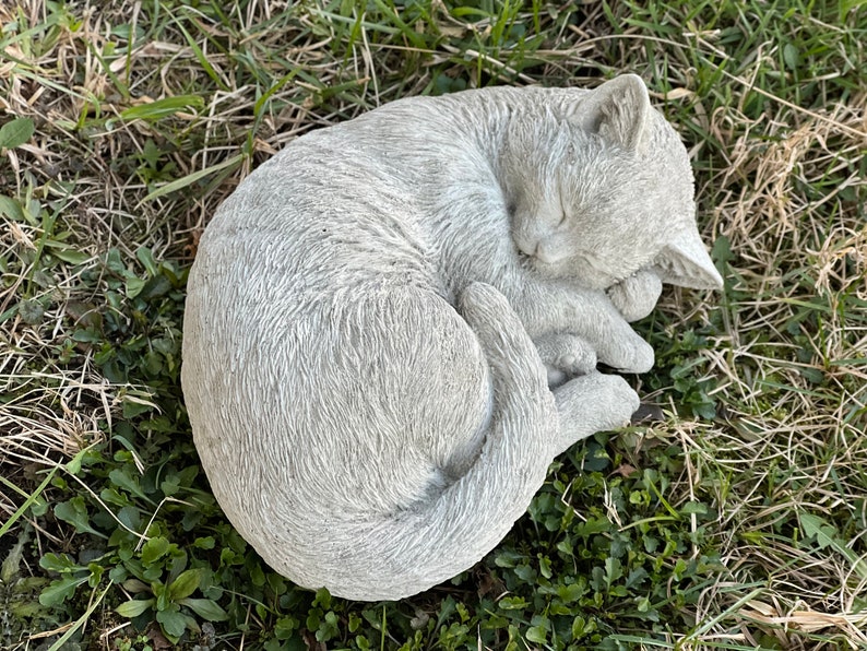 May include: A gray concrete garden statue of a cat curled up sleeping. The cat is lying on its side with its head resting on its paws. The statue is detailed with fur texture and realistic features.