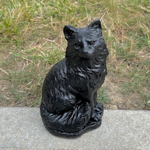 Peut inclure: Une figurine de chat en c&eacute;ramique noire pos&eacute;e sur une surface en b&eacute;ton gris. Le chat est assis avec sa queue enroul&eacute;e autour de son corps. Le chat a un visage d&eacute;taill&eacute; avec un long museau et de grandes oreilles.