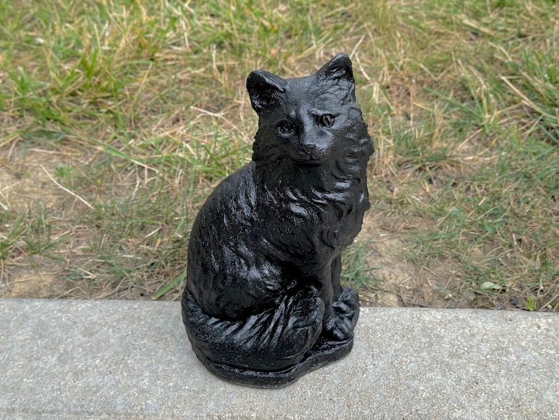 Peut inclure: Une figurine de chat en c&eacute;ramique noire pos&eacute;e sur une surface en b&eacute;ton gris. Le chat est assis avec sa queue enroul&eacute;e autour de son corps. Le chat a un visage d&eacute;taill&eacute; avec un long museau et de grandes oreilles.