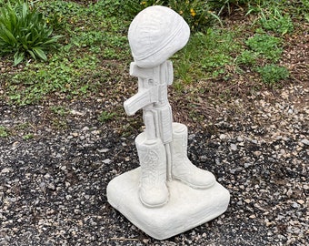 May include: White concrete statue of a fallen soldier. The statue depicts a helmet, rifle, and boots. The statue is on a square base.