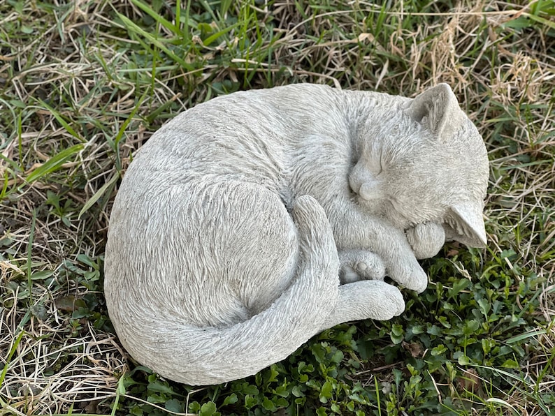 May include: A gray concrete garden statue of a cat curled up sleeping. The cat has a detailed face and fur texture.