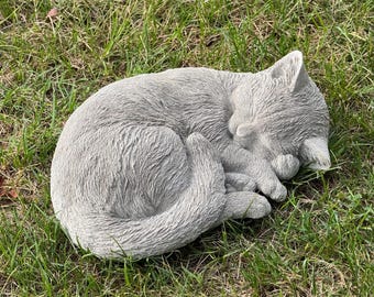 May include: A gray, sleeping cat statue curled up on a bed of green grass. The cat is in a relaxed pose, with its paws tucked in and its tail wrapped around its body. The statue is made of a textured material.