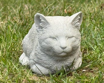 May include: A gray cat statue with closed eyes, resting in a relaxed pose. The cat sculpture is made of a textured material and is surrounded by green grass. A decorative item for cat lovers.