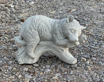 May include: A gray bear cub sculpture is resting on a log. The bear is lying down with its eyes closed, appearing to be asleep. The sculpture is made of a stone-like material and is placed outdoors.