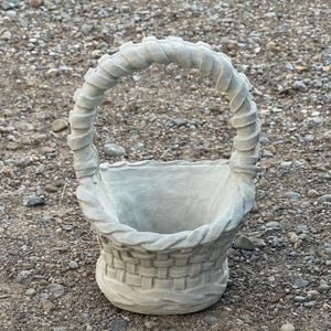 May include: A light gray, cement basket with a woven design and a curved handle. The basket has a round base and is suitable for holding plants or other decorative items.