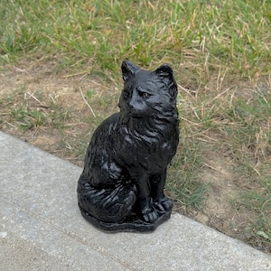 Peut inclure: Statue de chat noir avec une texture poilue. Le chat est assis avec sa queue enroul&eacute;e autour de ses pattes. La statue est faite d'un mat&eacute;riau sombre et solide.