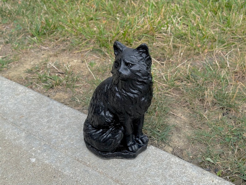 Peut inclure: Statue de chat noir avec une texture poilue. Le chat est assis avec sa queue enroul&eacute;e autour de ses pattes. La statue est faite d'un mat&eacute;riau sombre et solide.