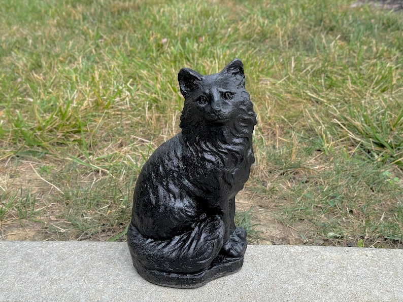 Peut inclure: Statue de chat noir en b&eacute;ton. Le chat est assis, la queue enroul&eacute;e autour de ses pattes. La statue est d&eacute;taill&eacute;e avec de la fourrure et des traits du visage.