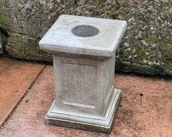 May include: A gray concrete pedestal with a square top and a round hole in the center. The pedestal is sitting on a concrete surface.