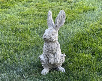 May include: A gray concrete rabbit statue with long ears, sitting upright on its hind legs. The rabbit has a textured surface and is set against a backdrop of green grass. A decorative garden statue.