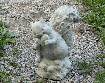 May include: A gray concrete squirrel statue holding a nut. The squirrel has a bushy tail and is sitting on a textured base. The statue is outdoors on a bed of small rocks.