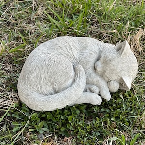 May include: A gray concrete sculpture of a cat curled up sleeping. The sculpture is detailed with fur and has a realistic appearance.