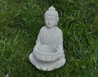 May include: A white concrete statue of a meditating Buddha holding a small bowl. The statue is sitting on a bed of green grass.