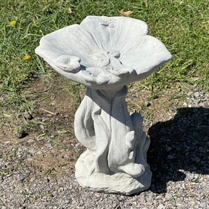 K&ouml;nnte beinhalten: Ein graues Vogelbad aus Beton in Form einer Blume mit einer breiten, flachen Schale und einem detaillierten Sockel. Der Sockel ist so gestaltet, dass er Bl&auml;tter und Stiele nachahmt.
