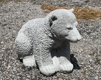 May include: A gray concrete sculpture of a bear cub sitting on its haunches. The sculpture is detailed with fur texture and has a realistic appearance.
