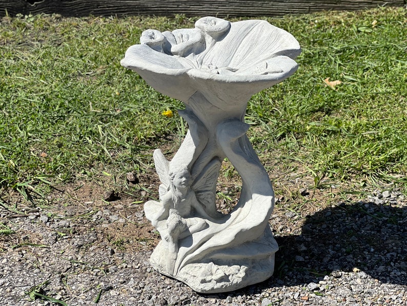 K&ouml;nnte beinhalten: Ein wei&szlig;er Beton-Vogelbad im Garten in Form einer Blume mit einer Fee, die auf dem Stiel sitzt. Das Vogelbad hat eine flache Schale f&uuml;r Wasser und ein dekoratives Design.