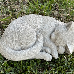 May include: A gray concrete garden statue of a cat curled up sleeping. The cat is lying on its side with its head resting on its paws. The statue is detailed with fur and a realistic sleeping expression.