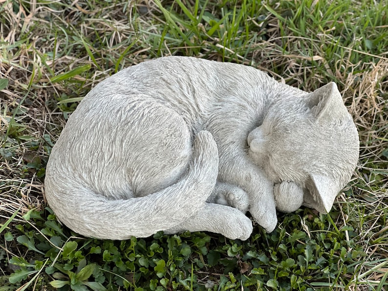 May include: A gray concrete garden statue of a cat curled up sleeping. The cat is lying on its side with its head resting on its paws. The statue is detailed with fur and a realistic sleeping expression.