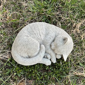 May include: A gray concrete cat statue curled up in a sleeping position. The statue is placed on a bed of green grass.