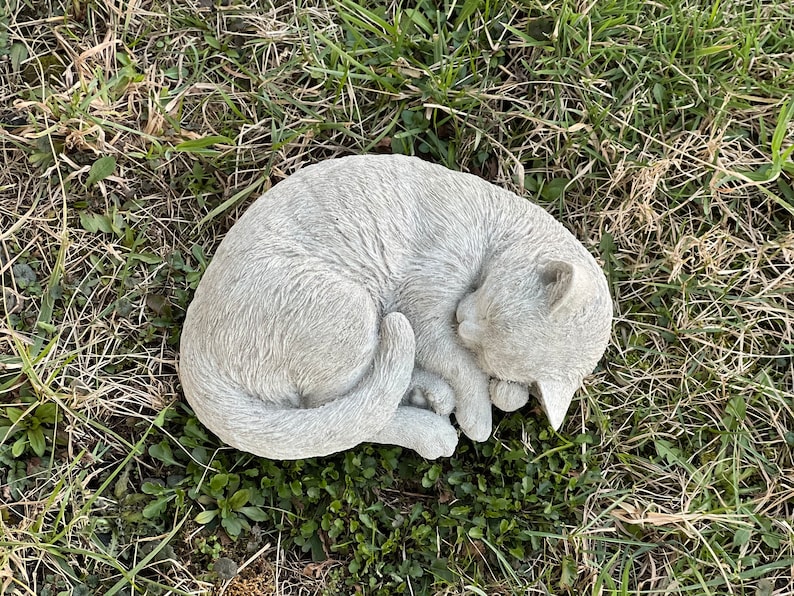 May include: A gray concrete cat statue curled up in a sleeping position. The statue is placed on a bed of green grass.
