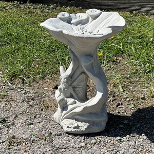 K&ouml;nnte beinhalten: Ein grauer Betonsteinvogelbad in Form einer Blume mit einer Fee, die auf einem Blatt sitzt. Das Vogelbad hat eine breite, flache Schale und einen hohen, schlanken Sockel.