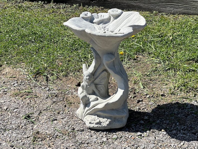 K&ouml;nnte beinhalten: Ein grauer Betonsteinvogelbad in Form einer Blume mit einer Fee, die auf einem Blatt sitzt. Das Vogelbad hat eine breite, flache Schale und einen hohen, schlanken Sockel.