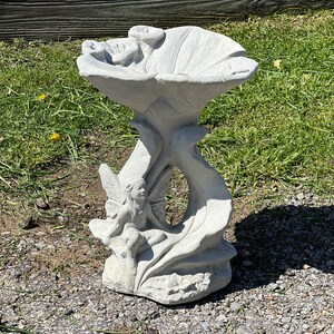 K&ouml;nnte beinhalten: Ein wei&szlig;er Vogelbad aus Beton in Form einer Blume mit einer Fee, die auf einem Blatt sitzt. Das Vogelbad hat eine breite, flache Schale und einen hohen, schlanken Stiel.