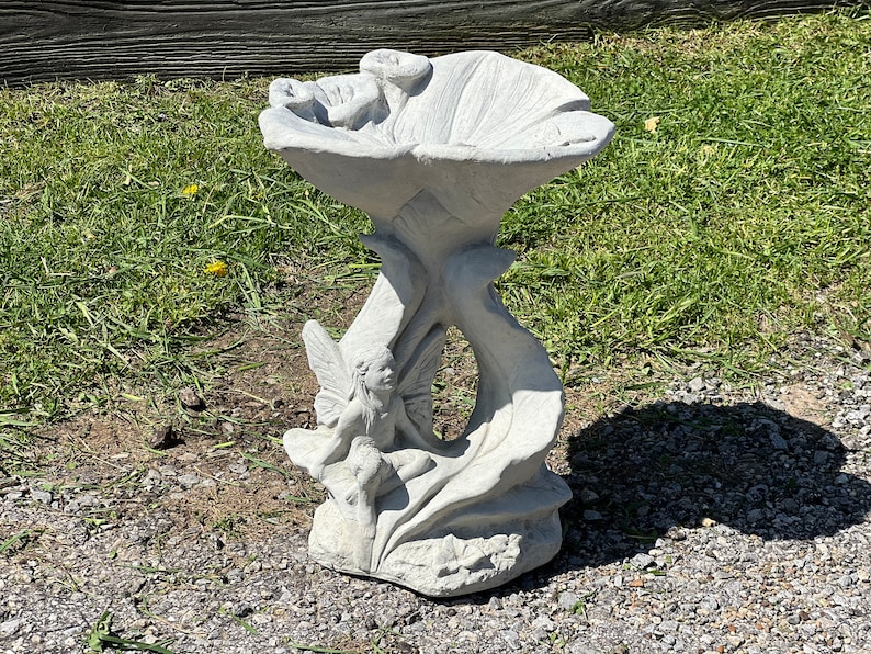 K&ouml;nnte beinhalten: Ein wei&szlig;er Vogelbad aus Beton in Form einer Blume mit einer Fee, die auf einem Blatt sitzt. Das Vogelbad hat eine breite, flache Schale und einen hohen, schlanken Stiel.