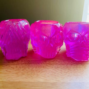 May include: Three vibrant pink skull-shaped candle holders. The translucent resin glows with a glittery effect, showcasing the detailed skull features. These decorative pieces are arranged on a wooden surface.