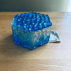 May include: A blue resin box with a crystal-like top. The box is square-shaped and has a textured surface. The box is approximately 3 inches wide and 3 inches tall. A one-dollar bill is next to the box.