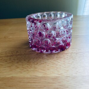 May include: A cylindrical resin candle holder featuring a clear, textured exterior with a repeating pattern of raised circular indentations. The interior and walls are embedded with small, vibrant magenta and deep purple dried flower petals.