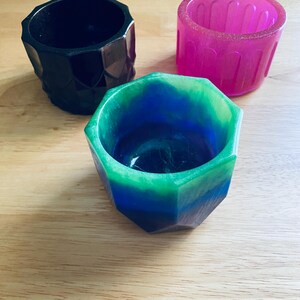 Puede incluir: Tres macetas de resina. Una es negra con un diseño geométrico, otra es rosa vibrante y la tercera es una mezcla de azul y verde. Las macetas son hexagonales y están sobre una superficie de madera.