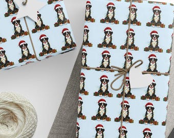 May include: Gift wrap featuring a repeating pattern of Bernese Mountain Dogs wearing Santa hats and holding presents on a light blue background. The wrapped gifts are tied with twine and adorned with white star-shaped tags. A ball of twine and scissors are also present.