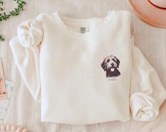 May include: A white sweatshirt featuring a black and white illustration of a dog with a pink bow on its head. The dog's name, "Katie", is printed beneath the illustration.
