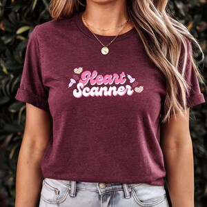 Puede incluir: Camiseta granate con mangas dobladas que muestra el texto "Heart Scanner" en rosa y blanco, con gráficos de corazón y ondas de sonido. La camiseta es de material suave, adecuada para uso casual.