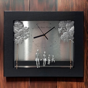 May include: A black framed clock with a silver face and silver hands. The clock face says "The Gaines Family". There are two silver trees on either side of the clock and a silver family of four sitting on a ledge below the clock.