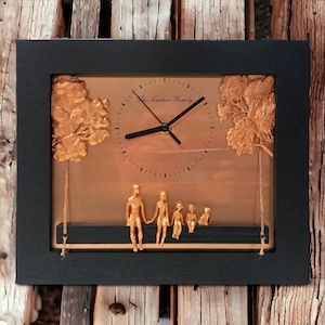 May include: A black framed clock with a copper background and gold accents. The clock face reads "The Sentinel Family".  The clock features a 3D design of a family sitting on a bench under two trees.