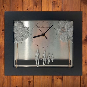 May include: A black framed wall clock with a silver face. The clock features a family of four figures sitting on a bench beneath two trees. The clock face reads "The Moy Family".
