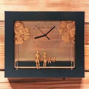 May include: A rectangular wall clock with a dark charcoal frame and a metallic gold face. The clock features black hands, a central dial, and the text The Hurst Family. Two gold tree reliefs flank the sides, and a gold family figurine sits on a dark horizontal bar suspended by thin strings.
