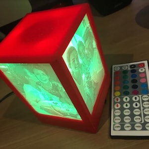 May include: A red cube-shaped lamp with a photo of three people inside. The lamp is illuminated with a green light and has a remote control with buttons for different light settings. The buttons on the remote control include: Quick, Slow, Auto, Flash, DIY1, DIY2, DIY3, DIY4, DIY5, DIY6, Jump1, Jump2, Fade1, Fade2.