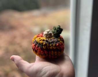 May include: A small crocheted pumpkin with brown, orange, yellow, and green yarn. The pumpkin is sitting in the palm of a hand.