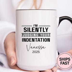 May include: White ceramic mug with a black handle and rim. The mug features the text "I'M SILENTLY JUDGING YOUR INDENTATION" in black, with the name "Vanessa" and the year "2025" below. The mug is held by a person.
