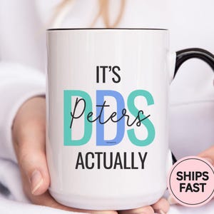 May include: White ceramic mug with black handle and rim, featuring the text "IT'S DDS Peters ACTUALLY" in teal, blue, and black. The mug is held by a person. A pink circle with "SHIPS FAST" is on the right.