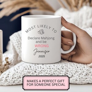 May include: White ceramic mug with the text "MOST LIKELY TO Declare Mahjong and be WRONG Jennifer 2025". The mug has a handle and is held by a person. A pink and black sign at the bottom says "MAKES A PERFECT GIFT FOR SOMEONE SPECIAL".