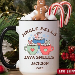 May include: A white ceramic mug with a black handle, featuring the text "Jingle Bells and Java Smells" and a personalized name and year. The mug is decorated with cartoon cocoa mugs, marshmallows, a gingerbread man, and candy canes.